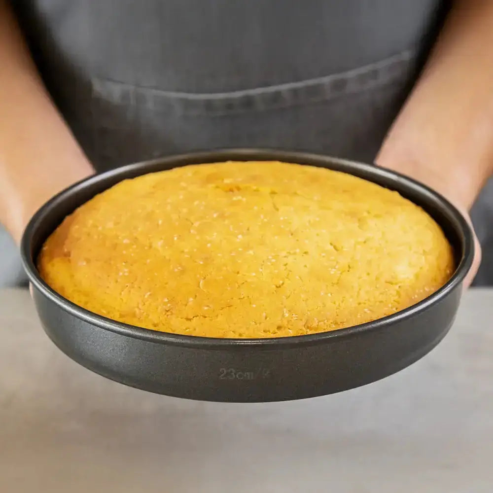 Baked cake in a round metal baking pan held by a person wearing an apron.