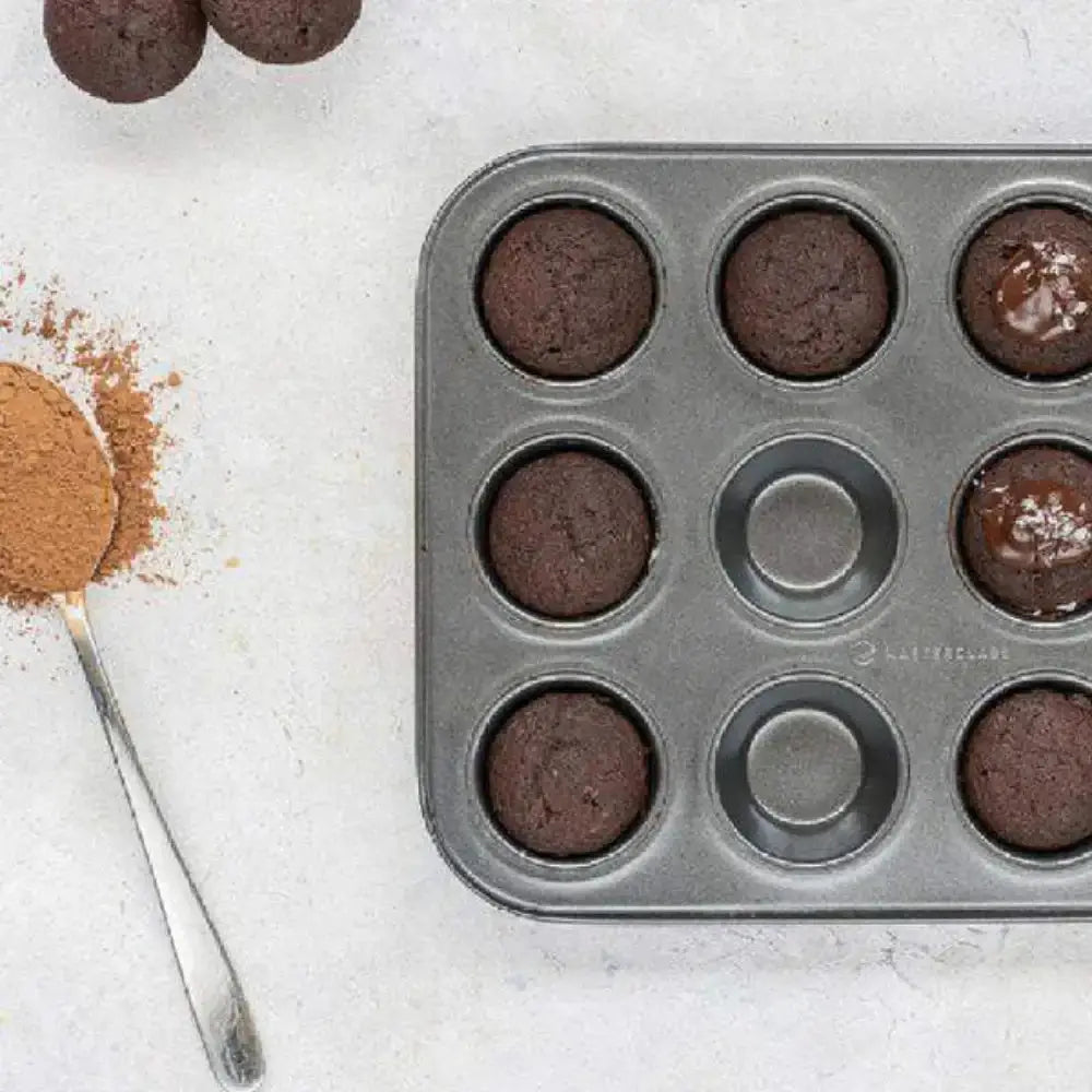 Muffin tin with chocolate muffins, cinnamon, and a spoon on a light surface