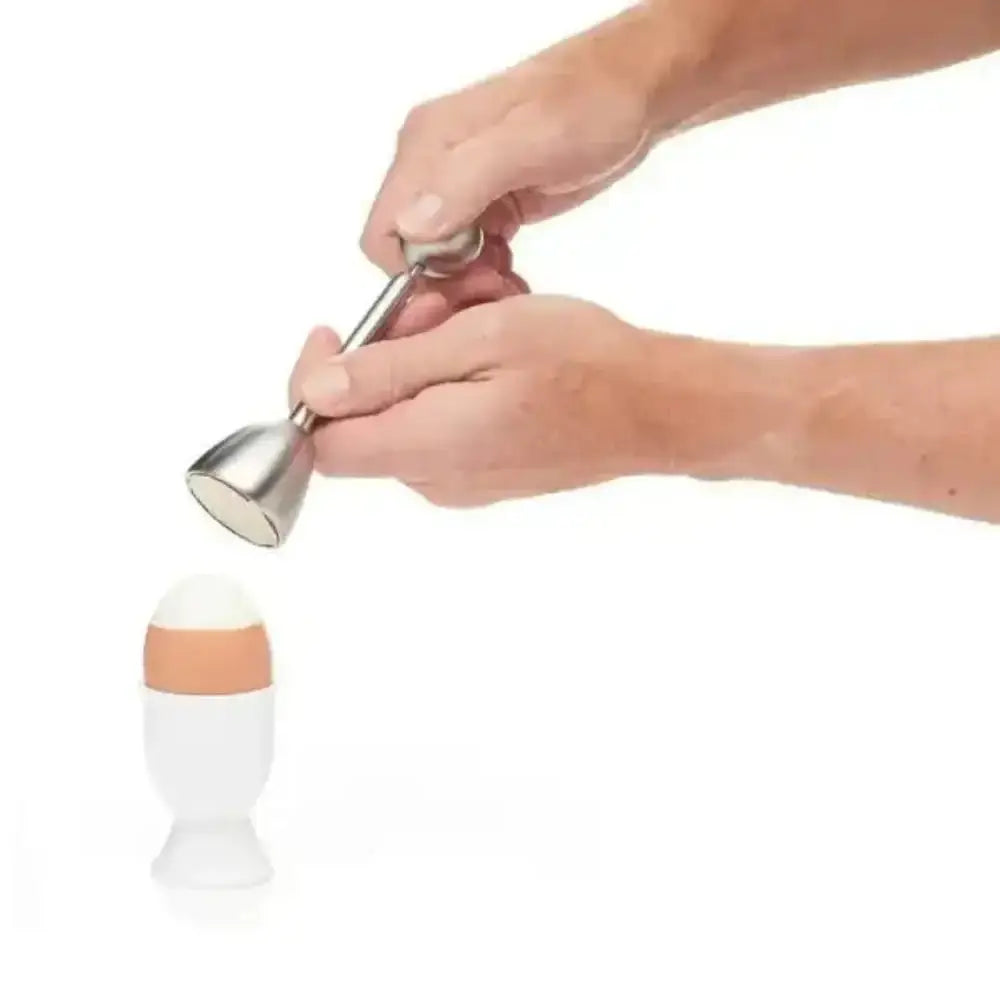 Person using a metal topper beater on an egg in a white holder against a white background