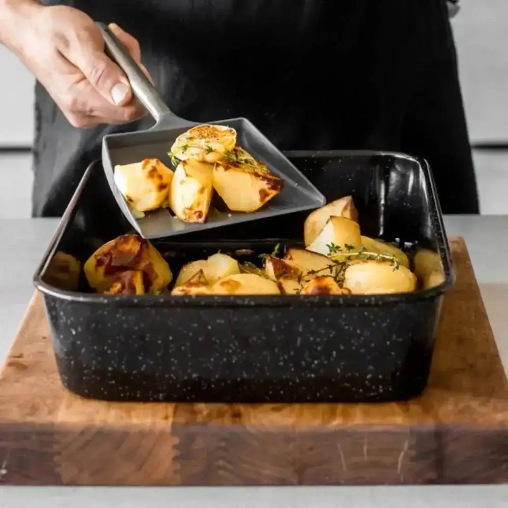 Person serving roasted potatoes from a black speckled roasting pan onto a square plate.