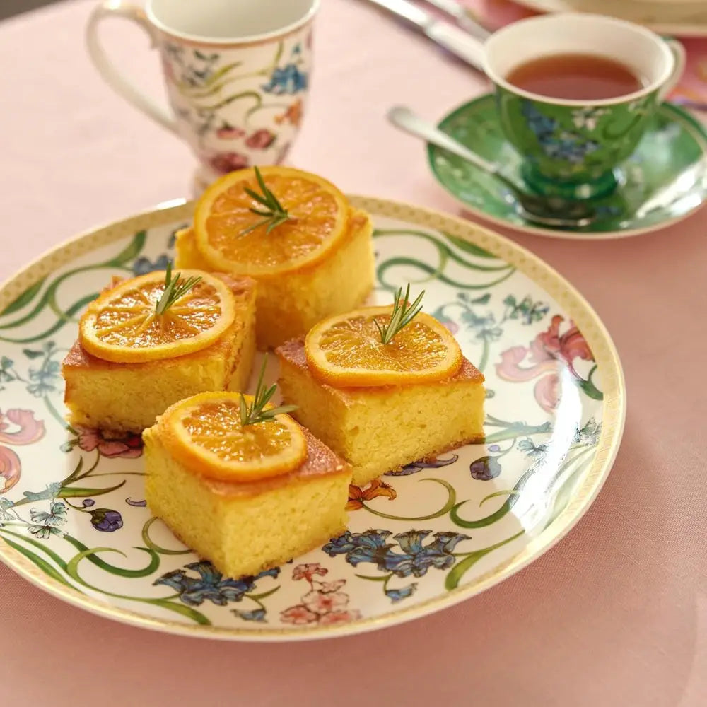 Slices of cake with orange slices and rosemary on a floral plate with tea cups in the background.