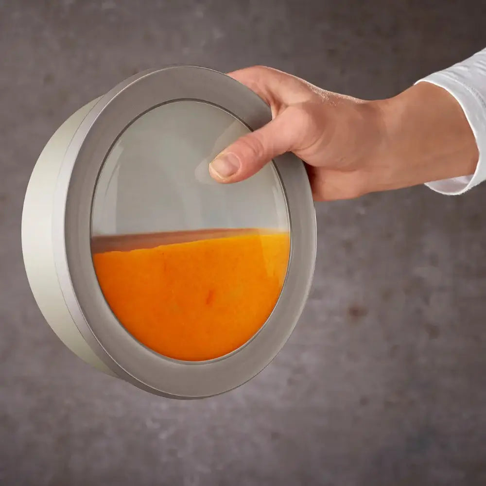 Hand holding a round container with orange liquid against a gray background