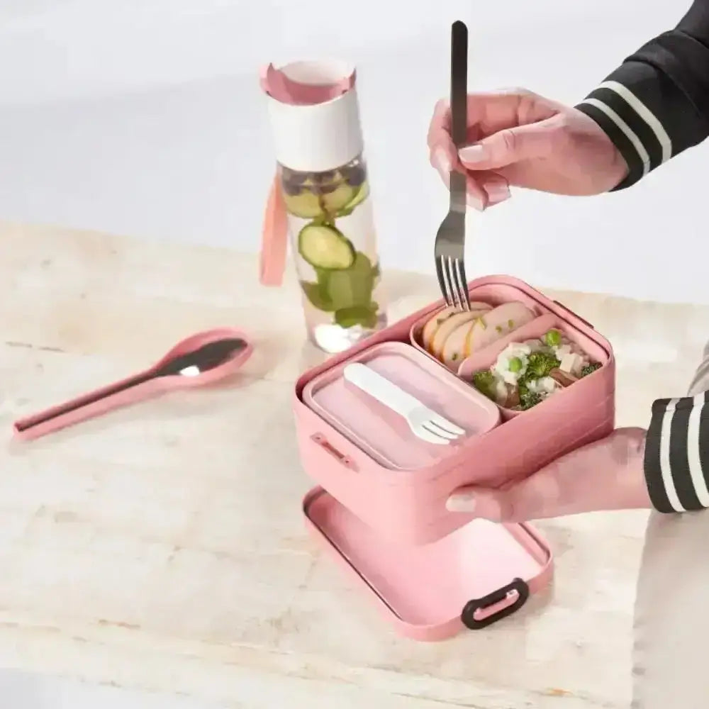 Pink lunch box with compartments, fork, and water bottle, held on a light wooden table