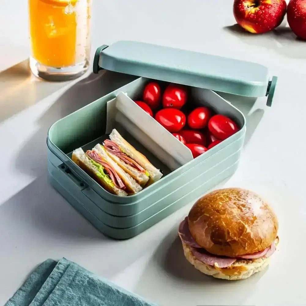 Green lunch box with compartments containing sandwiches and tomatoes, next to a burger and glass of juice on a white surface.