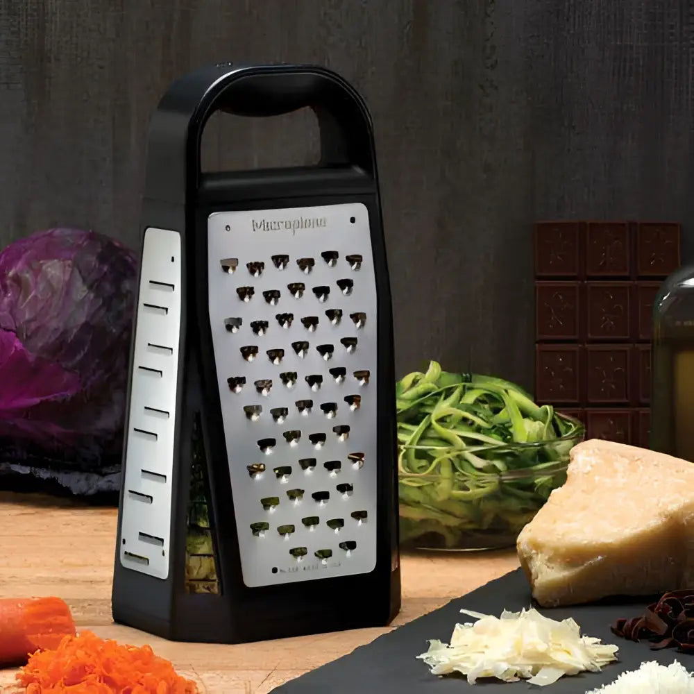 Box grater on a wooden surface with vegetables and cheese in the background