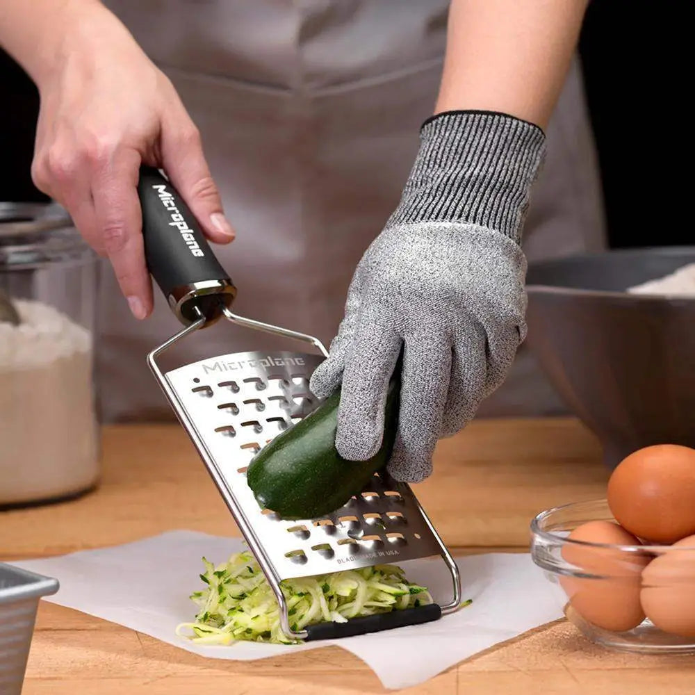 Person using a Microplane zester to shred a zucchini with a focus on safety with a cut-resistant glove.