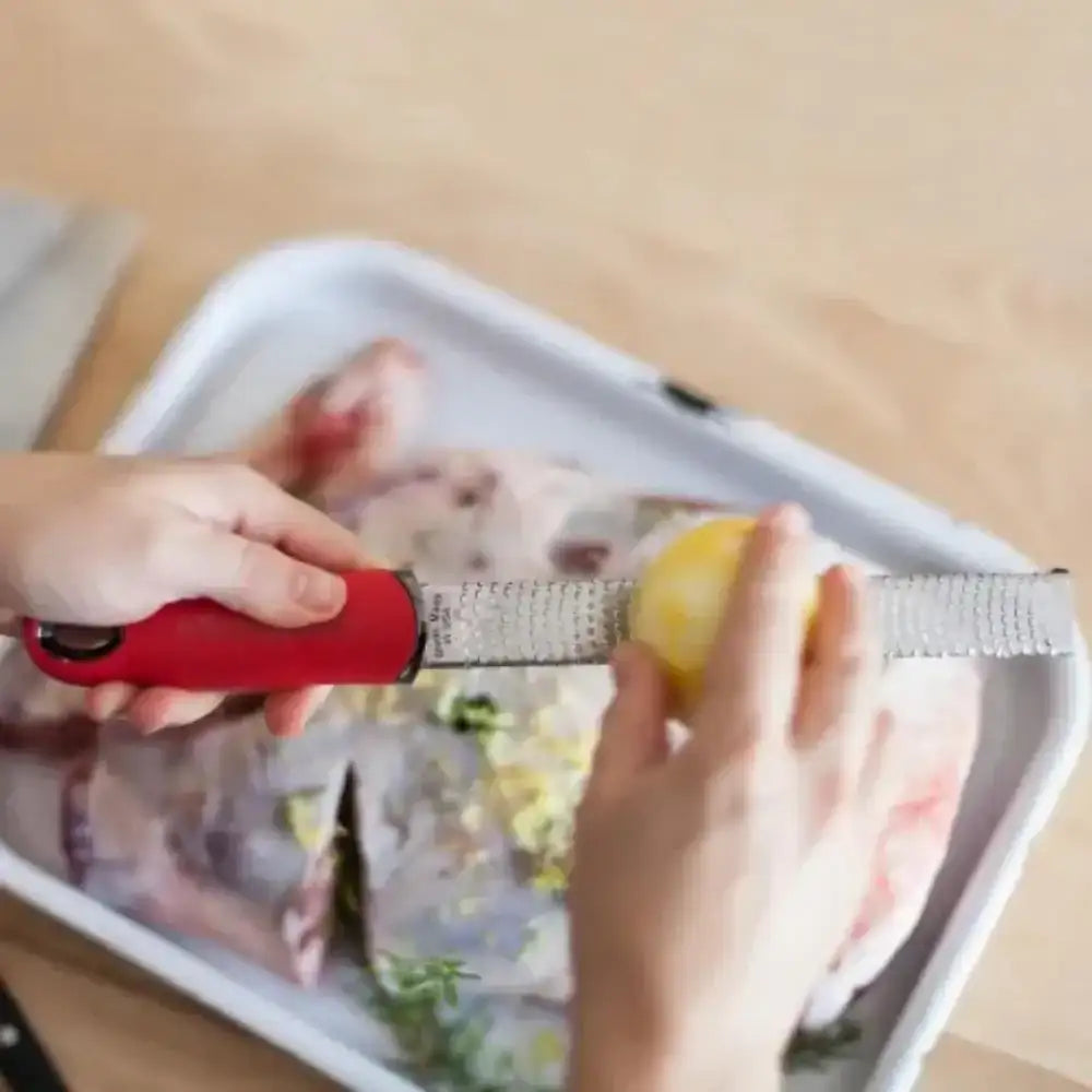 Person using a red microplane zester on a lemon over a white tray with a blurred background.