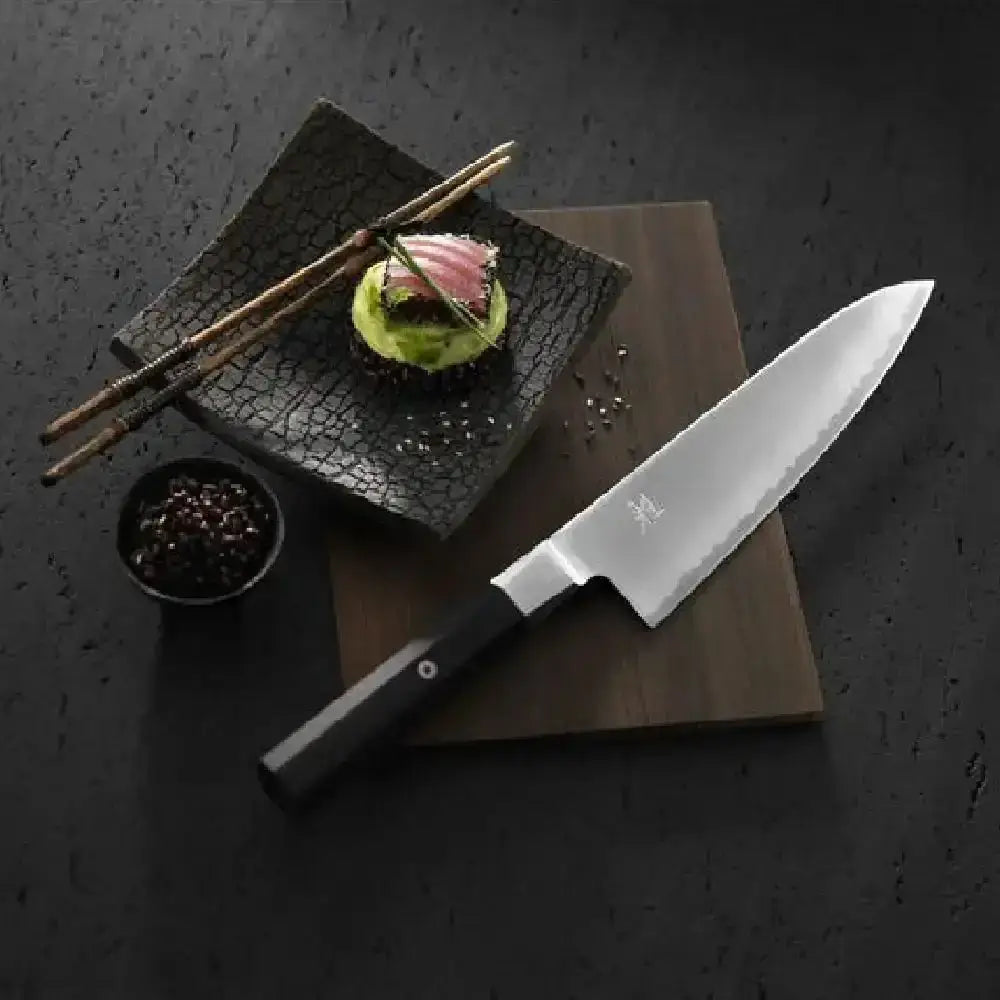 Chef's knife on a dark surface with a plate of food and chopsticks.