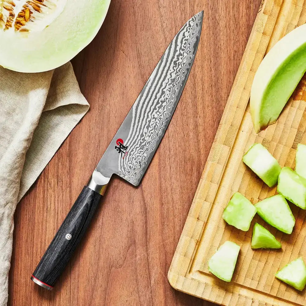 Chef's knife on a wooden surface with chopped melon and a cutting board.