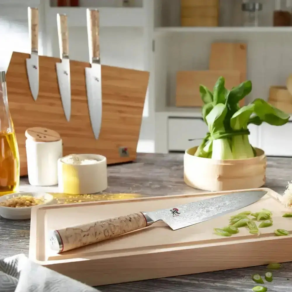 Kitchen scene with knives, cutting board, and vegetables on a wooden surface.