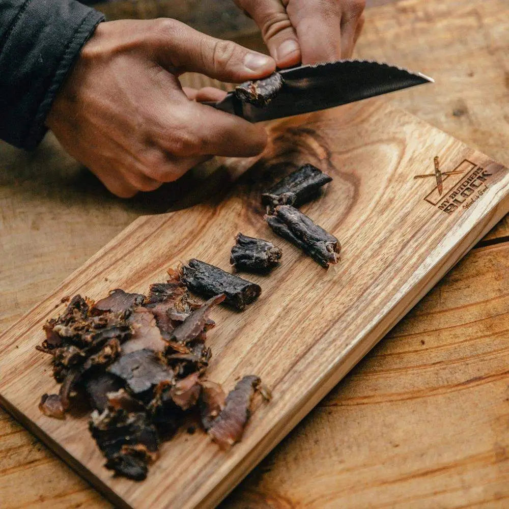 Person cutting dried meat on a wooden cutting board with a knife.