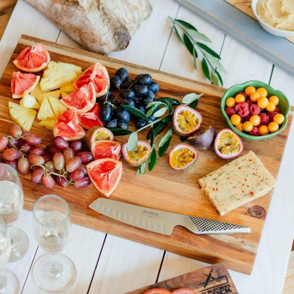 Wooden cutting board with fruits, cheese, and a knife on a white surface.