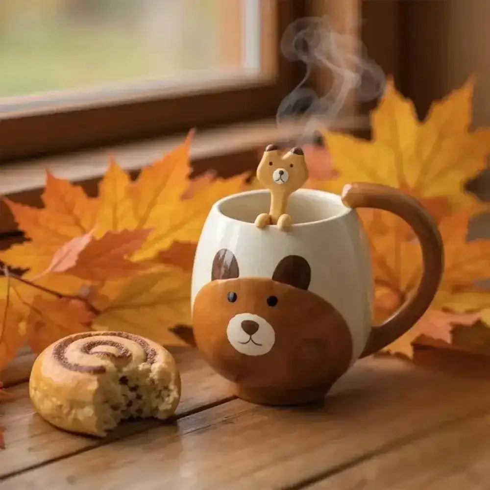 Ceramic mug with bear design, steaming cat-shaped object, and pastry on a wooden surface with autumn leaves.