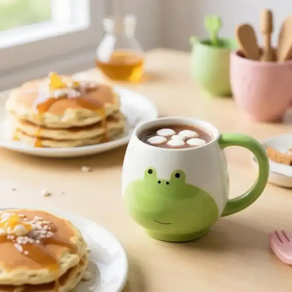 Green frog-shaped mug with hot chocolate and marshmallows on a table with pancakes and kitchen items.