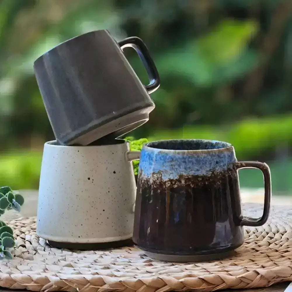 Three ceramic mugs stacked on a woven mat with a blurred green background