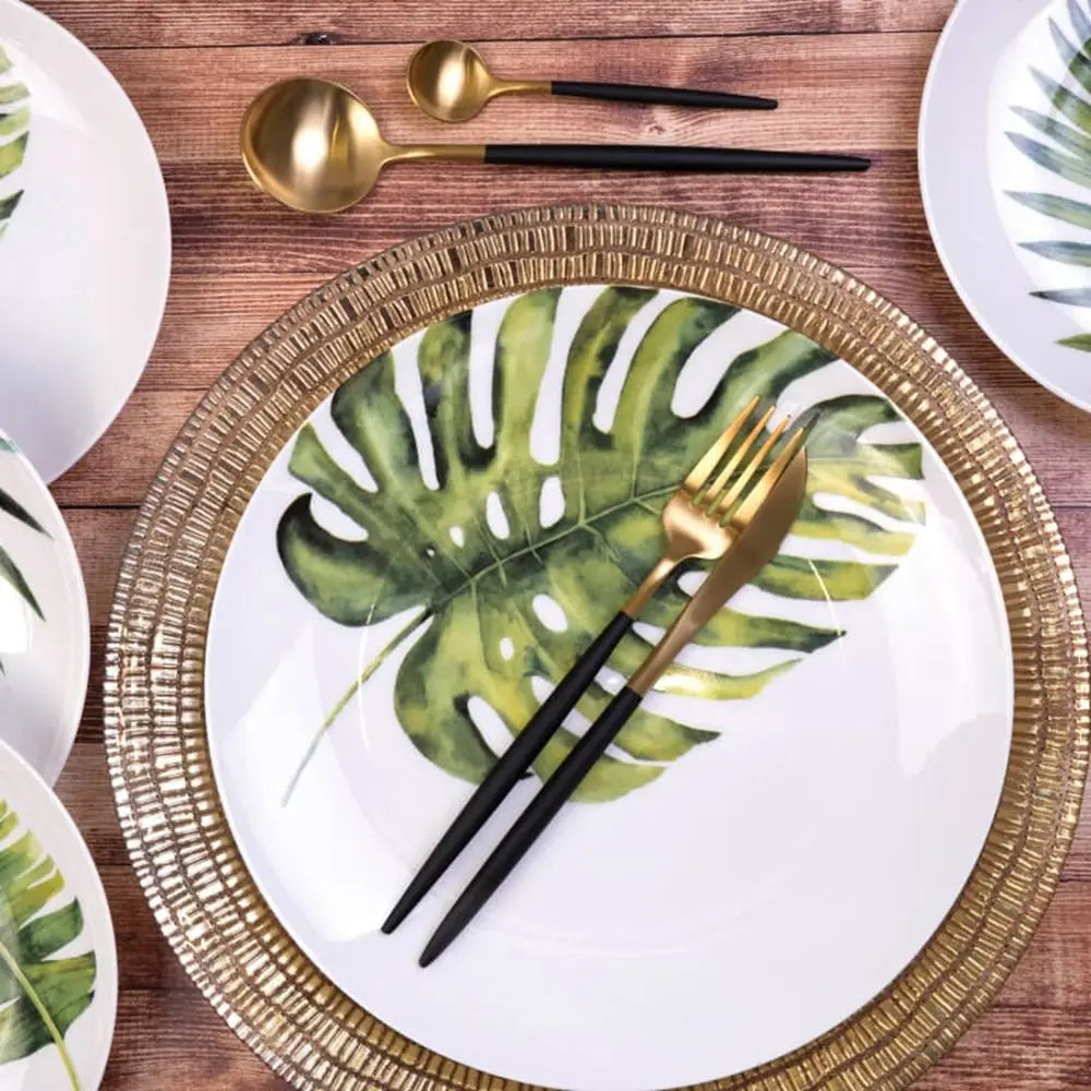 Table setting with tropical leaf-patterned plates, gold and black cutlery on a wooden table.