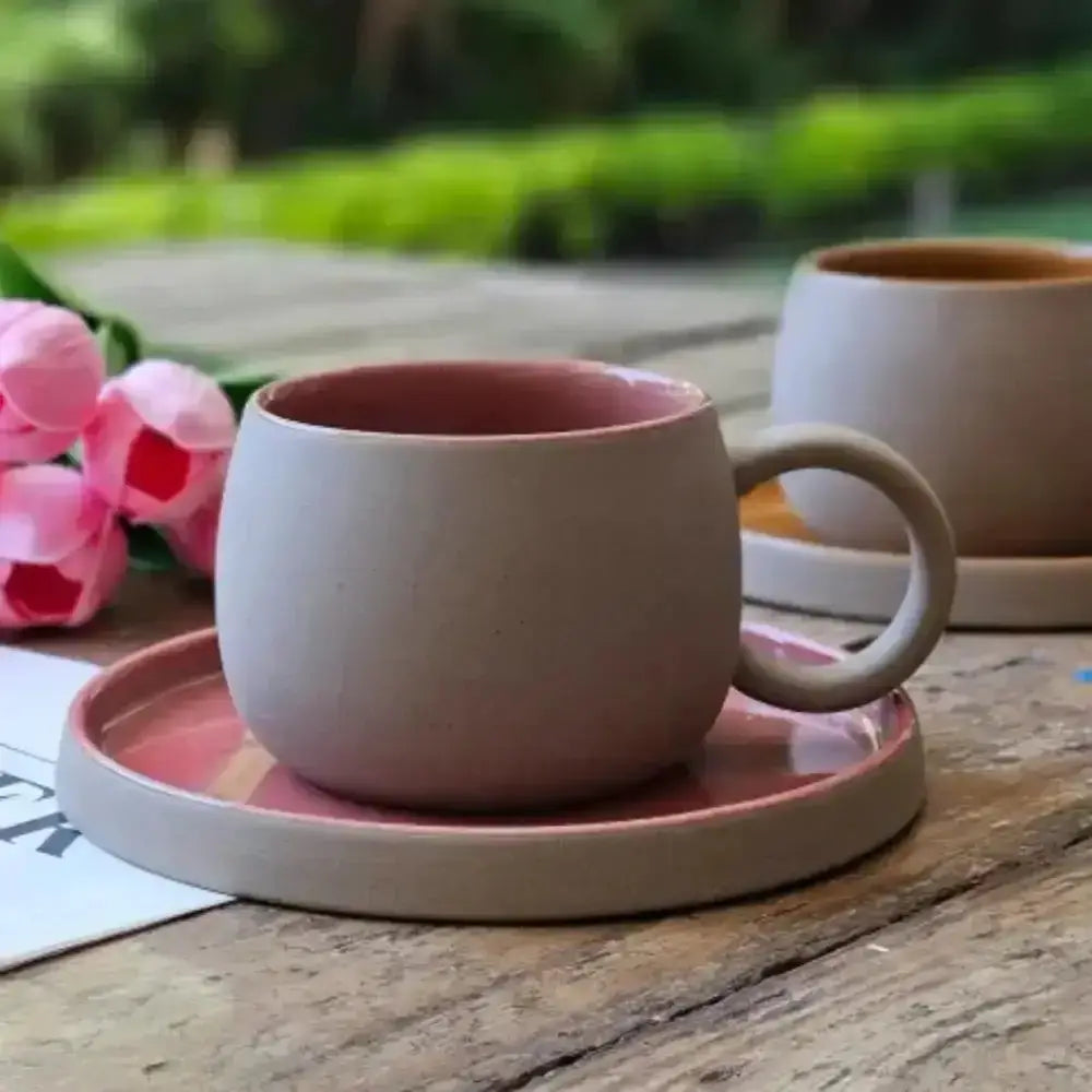 Pink ceramic cup and saucer on a wooden table with pink tulips and a plant in the background.