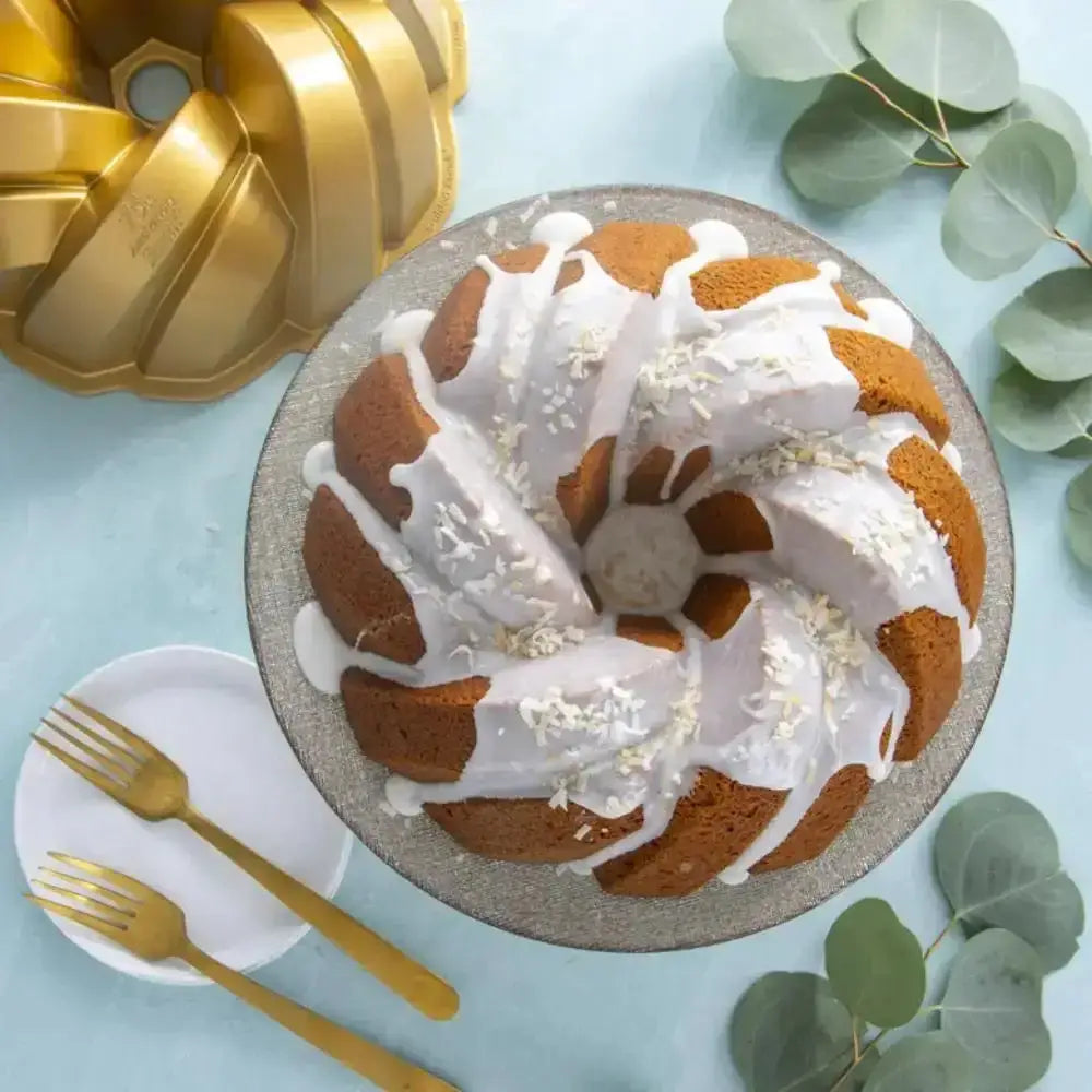 Bundt cake with white icing on a plate with gold cake pan and forks on a light blue background