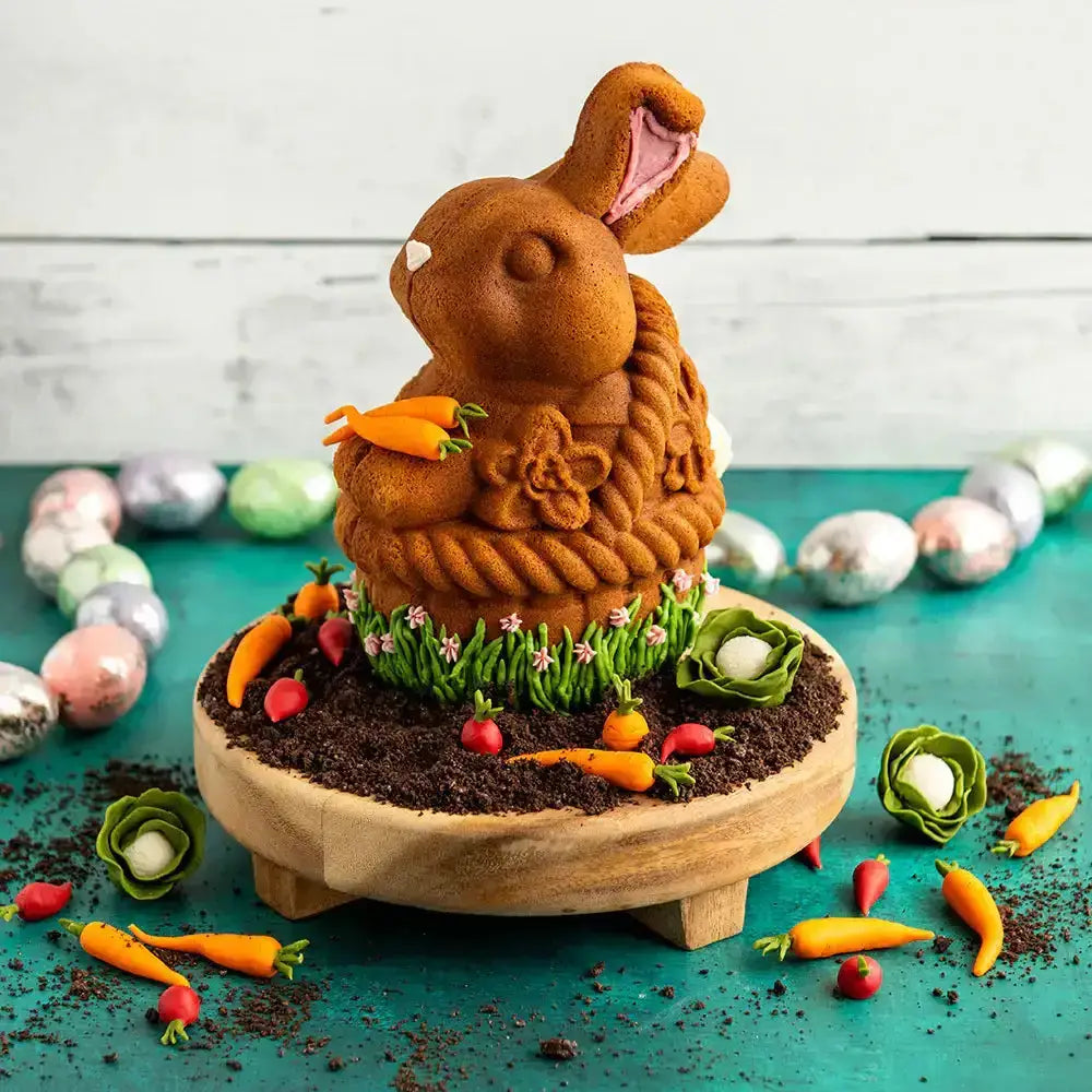 Decorative Easter bunny shaped cake with a wooden base, surrounded by Easter eggs and carrots on a teal surface.