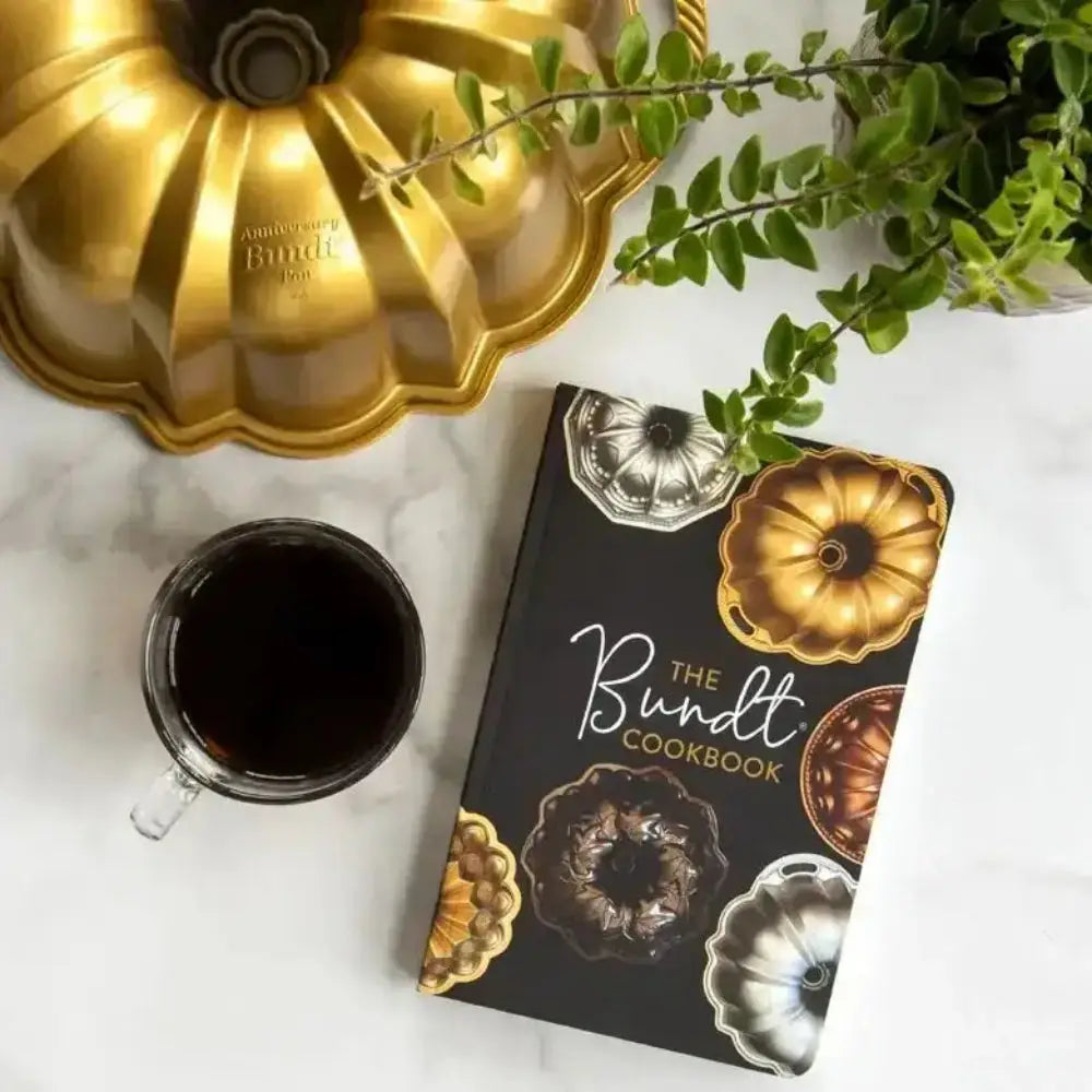 Bundt cake pan, coffee cup, and 'The Bundt Cookbook' on a marble surface with greenery.