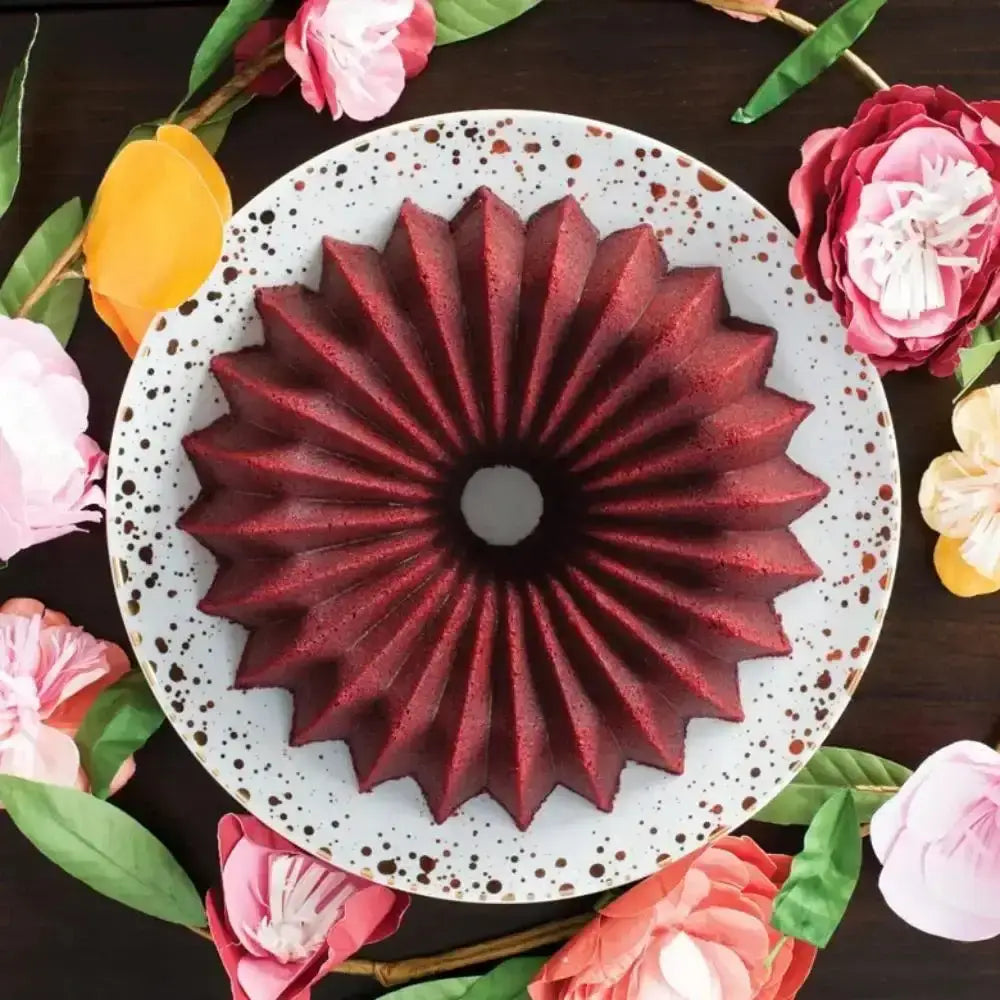 Red floral cake on a white plate surrounded by colorful flowers on a dark surface