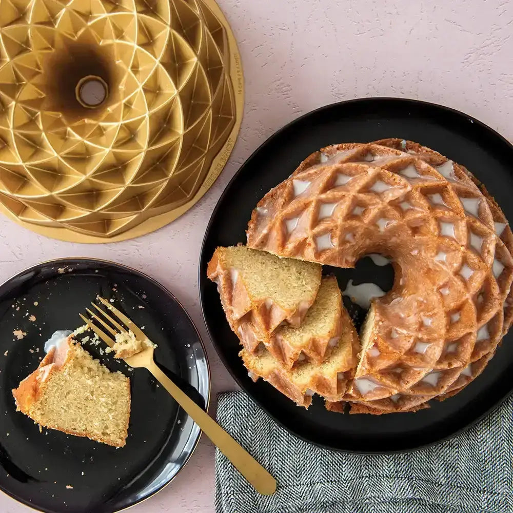Bundt cake with a slice on a black plate, gold cake pan, and gold fork on a light pink background.