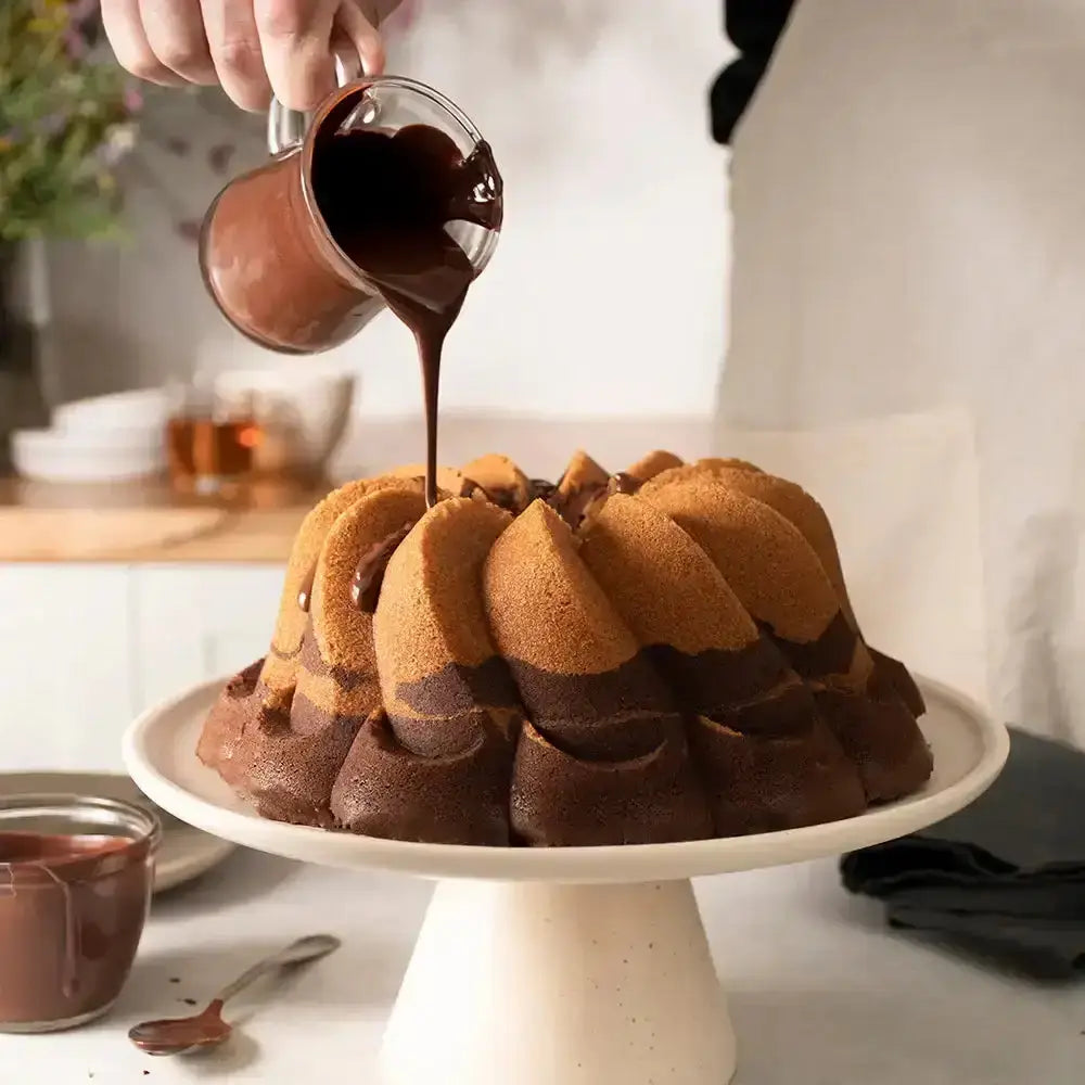 Chocolate sauce being poured over a bundt cake on a white stand.