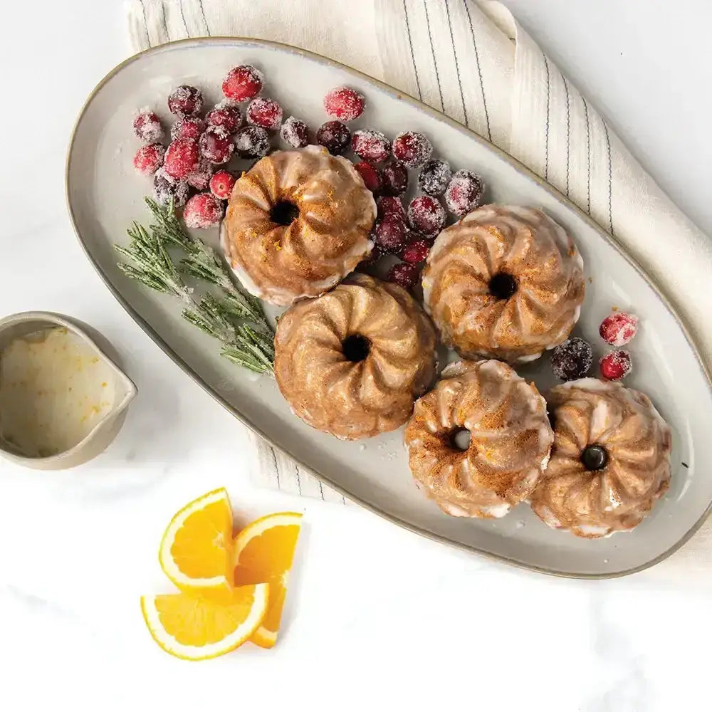 Four small Bundt cakes on a platter with frosted berries and orange slices on a white background