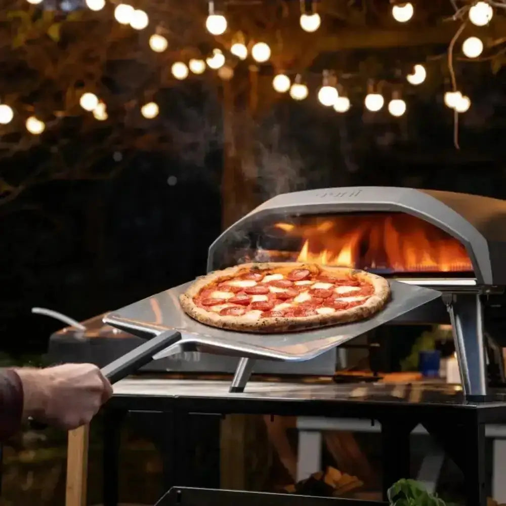 Person placing a pizza into an outdoor pizza oven with string lights in the background