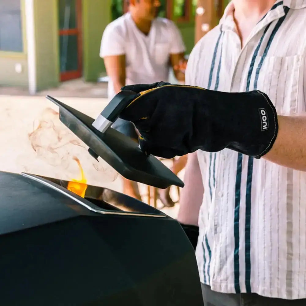 Person wearing a black glove using an outdoor oven, with another person in the background.