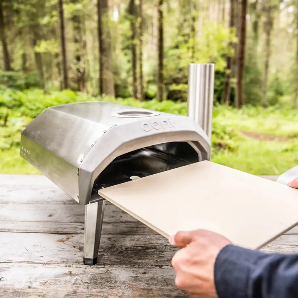 Portable pizza oven on a wooden surface with a person holding a pizza stone outdoors.