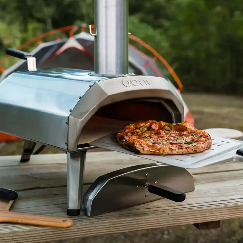 Outdoor pizza oven with a pizza on a wooden table, with a blurred background of greenery and a bicycle.
