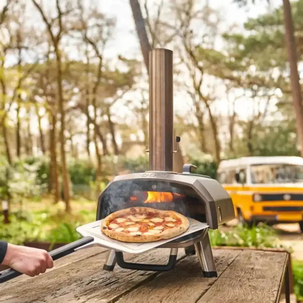 Portable pizza oven with a pizza being cooked outdoors, with a van in the background.