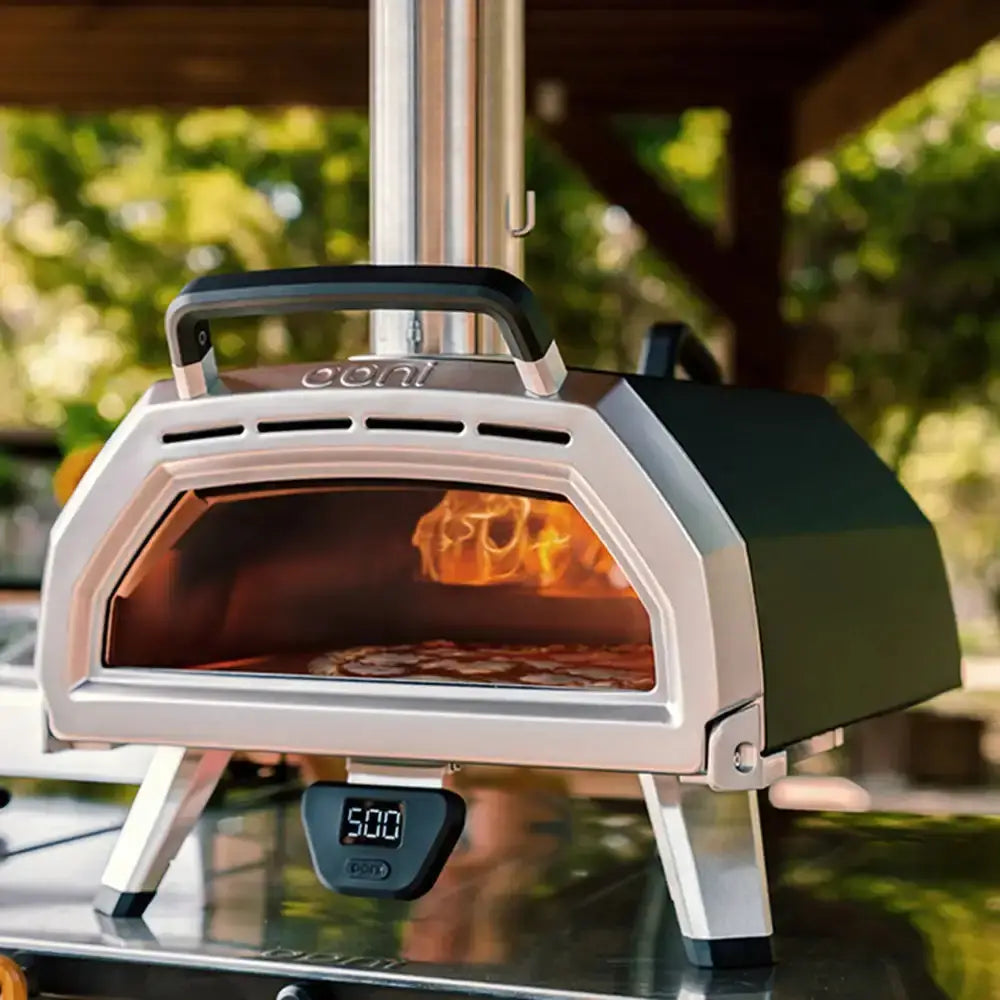 Portable outdoor pizza oven with flames inside, set against a blurred natural background.