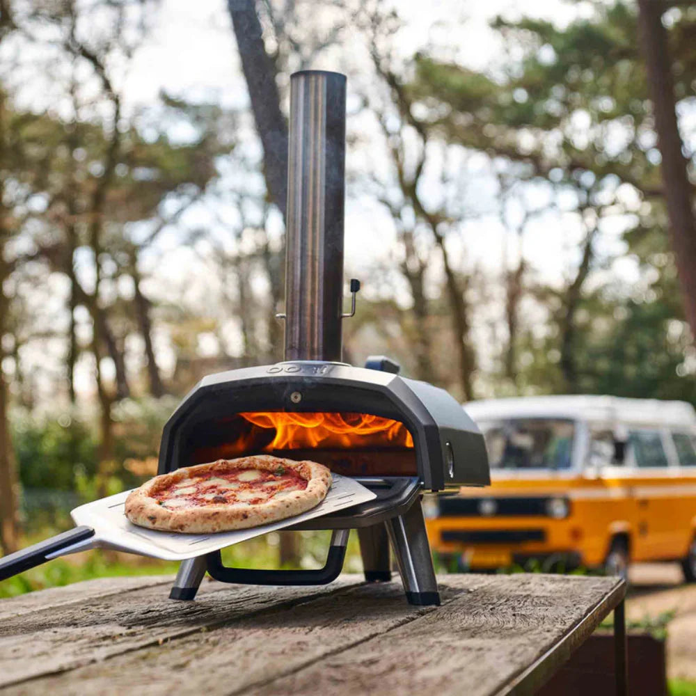 Portable pizza oven with a pizza inside, set outdoors with a blurred background of trees and a van.