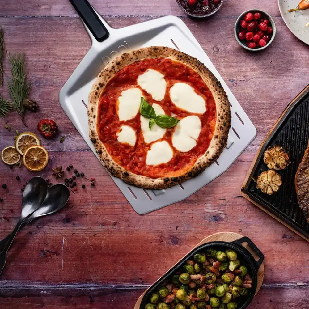 Wooden table with a pizza on a white pan, surrounded by various ingredients and dishes.
