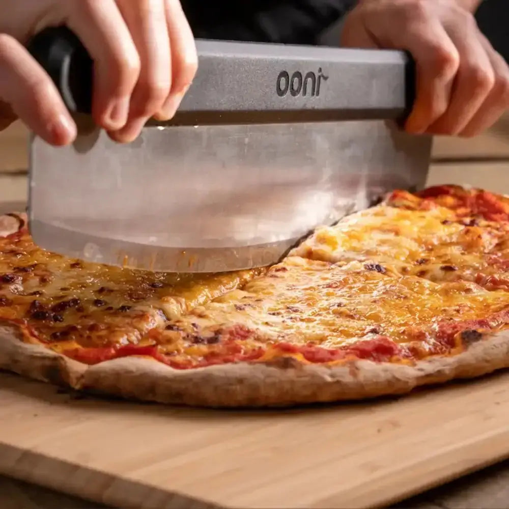 Person using a pizza cutter labeled 'Ooni' to slice a pizza on a wooden board.