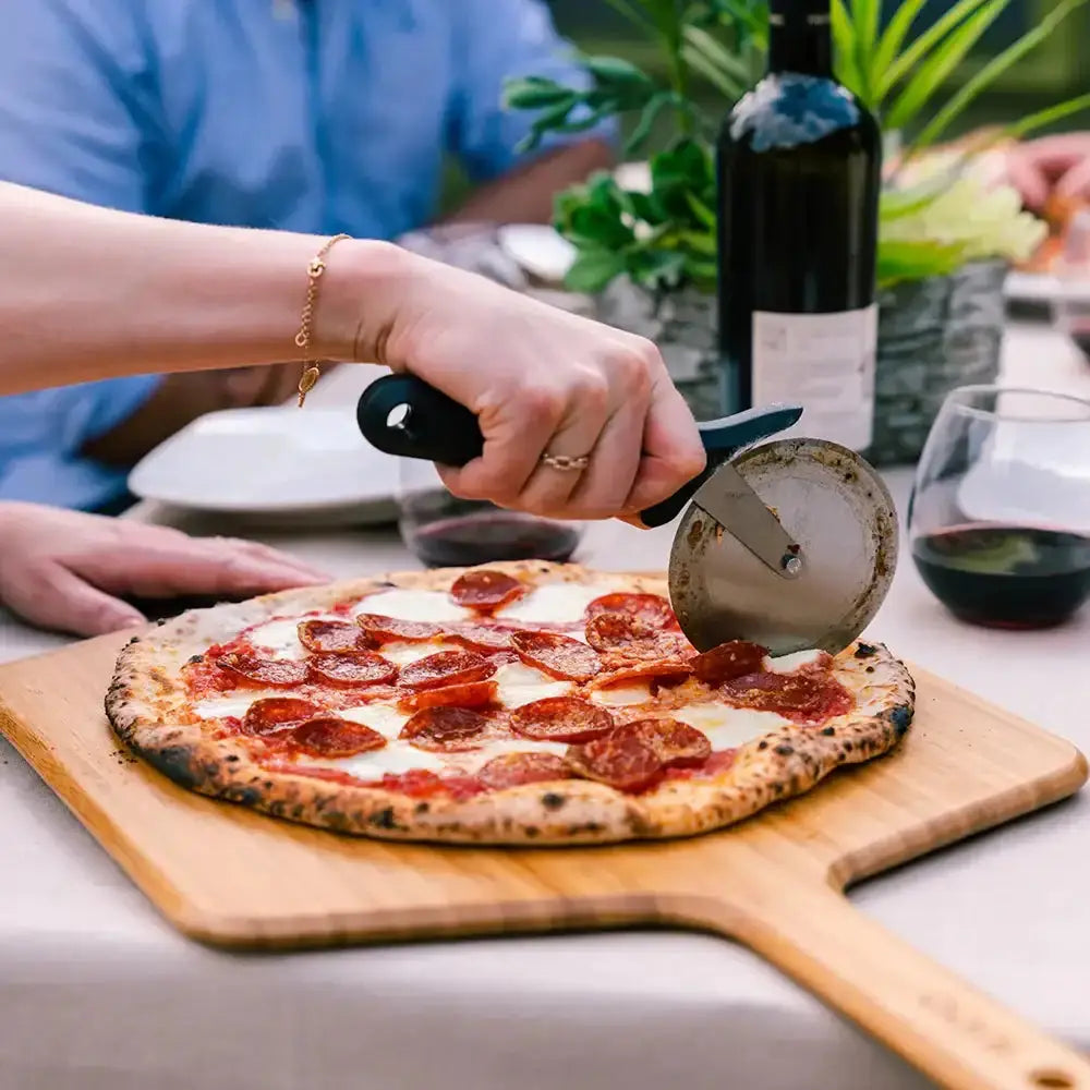 Person cutting a pepperoni pizza on a wooden board with a pizza cutter, surrounded by wine and plants.