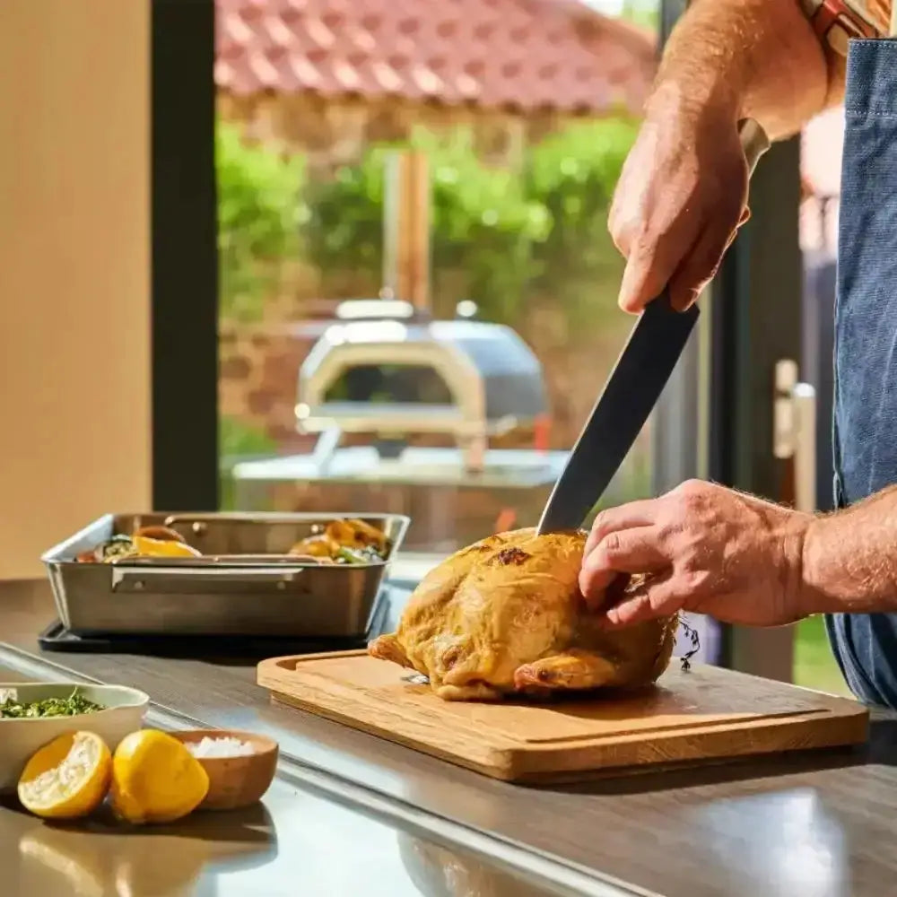 Person cutting a roasted chicken on a wooden cutting board with a view of a barbecue grill outside.