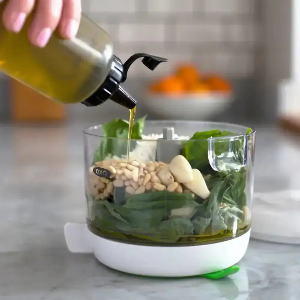 Hand pouring oil into a food processor with ingredients on a kitchen counter