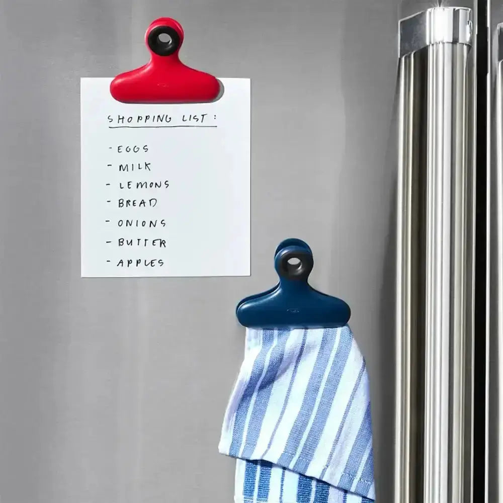 Red and blue magnetic clips holding a shopping list and a striped towel on a refrigerator.