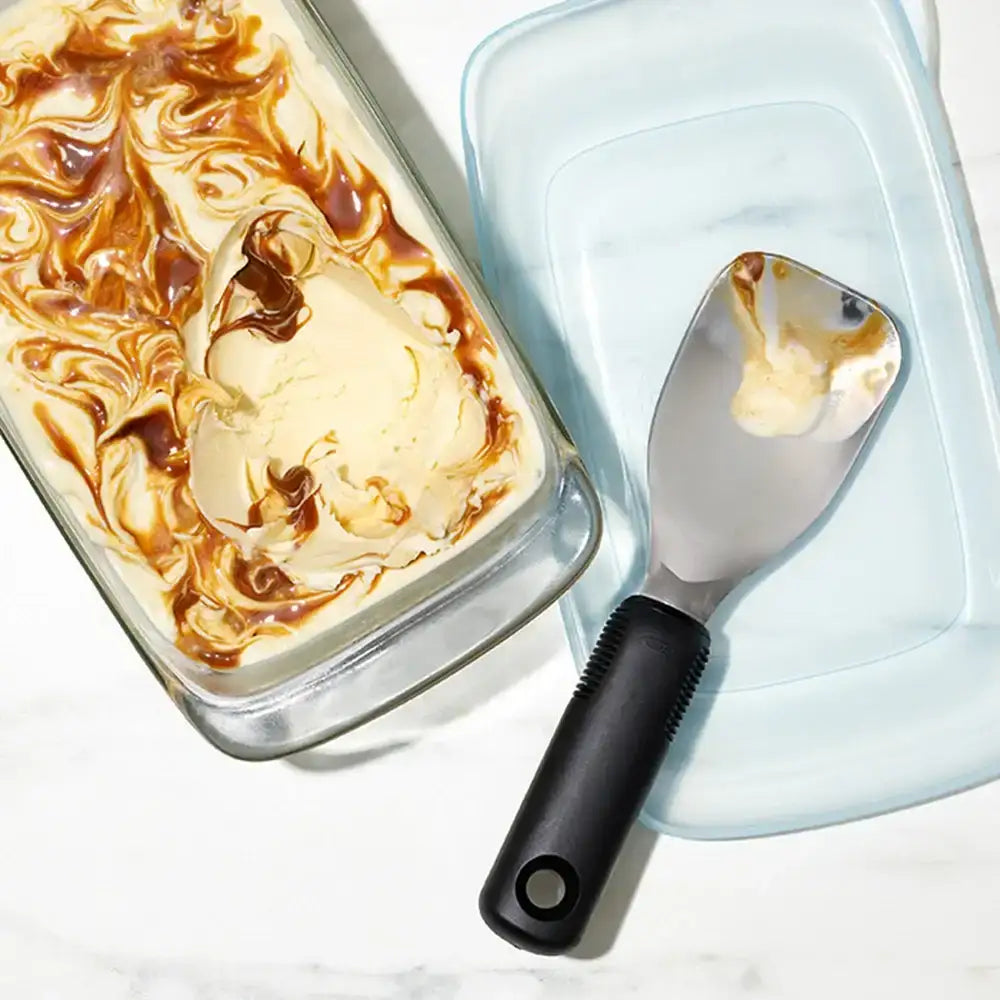 Glass container of ice cream with a swirl of caramel, black-handled spatula on a white background