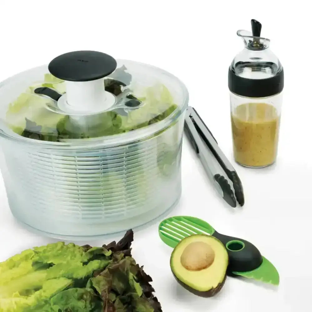 Salad spinner with lettuce, avocado, tongs, and a dressing container on a white background