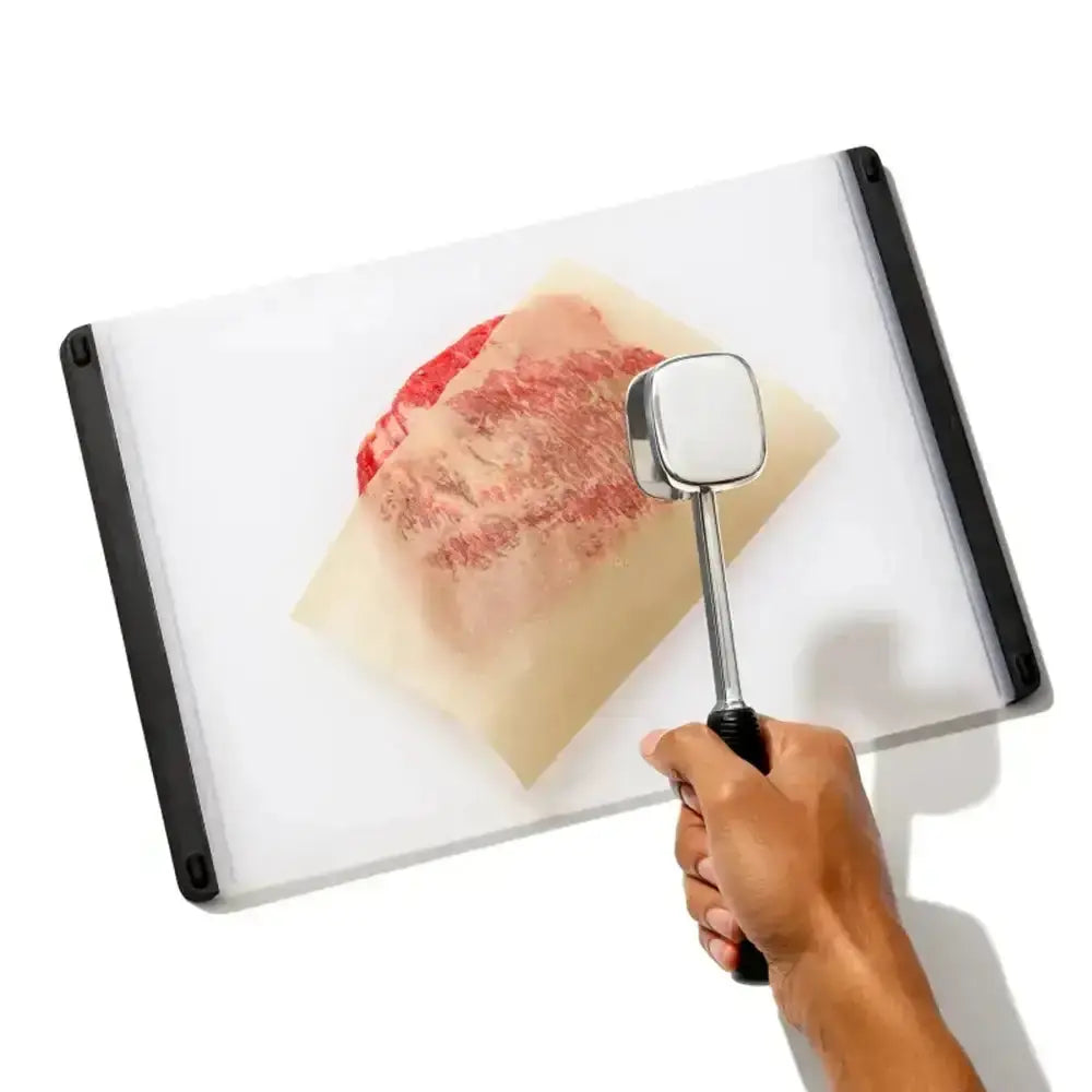 Hand holding a meat tenderizer over a cutting board with raw meat on a white background