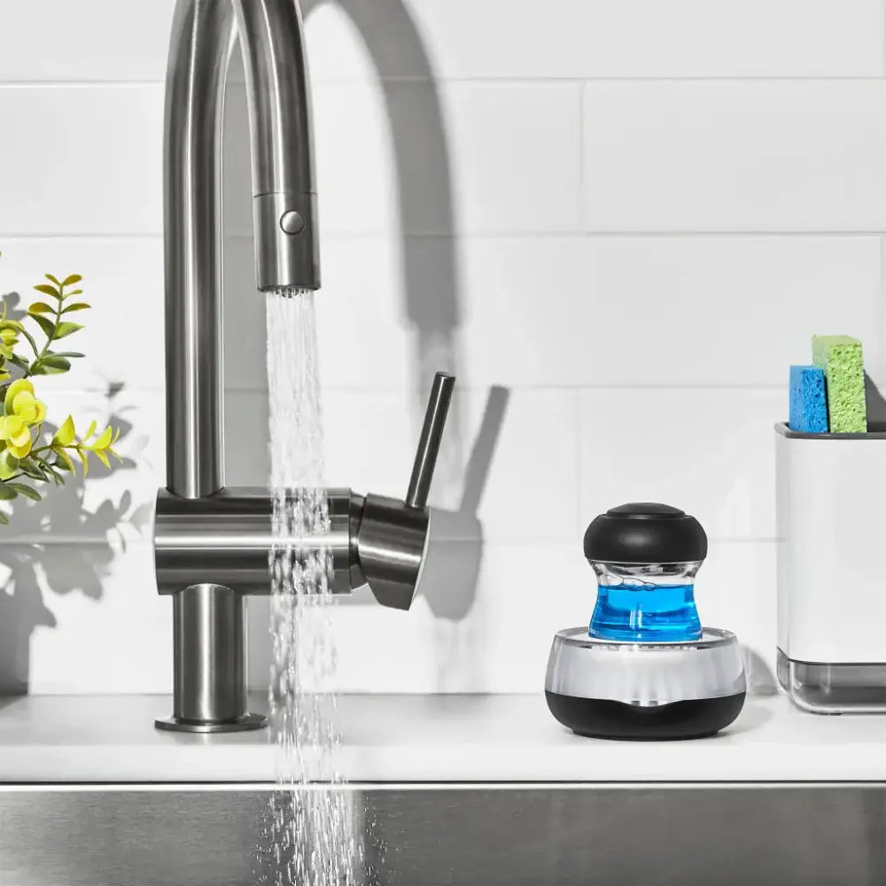 Oxo palm cleaning brush next to Kitchen faucet with water flowing, next to a black and blue faucet filter on a white countertop.