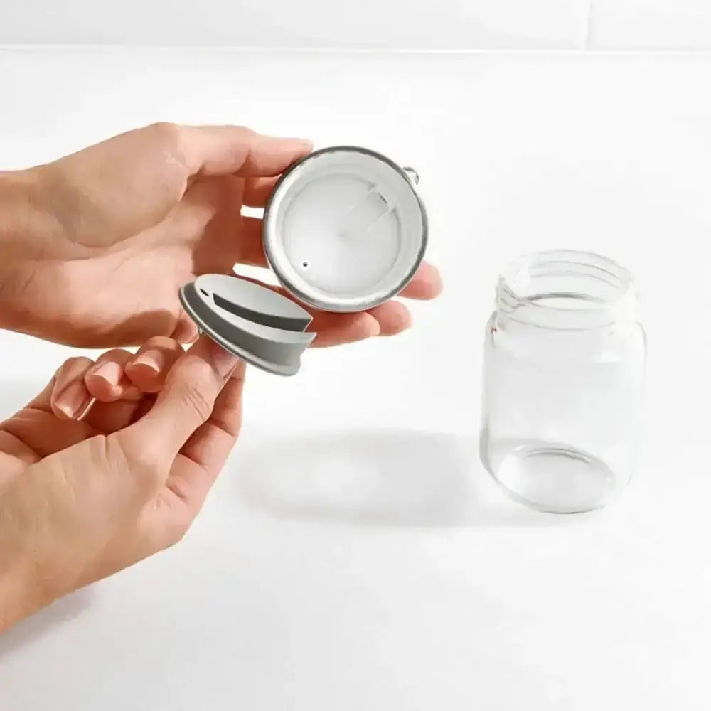 Person holding a metal infuser over a clear glass jar on a white background