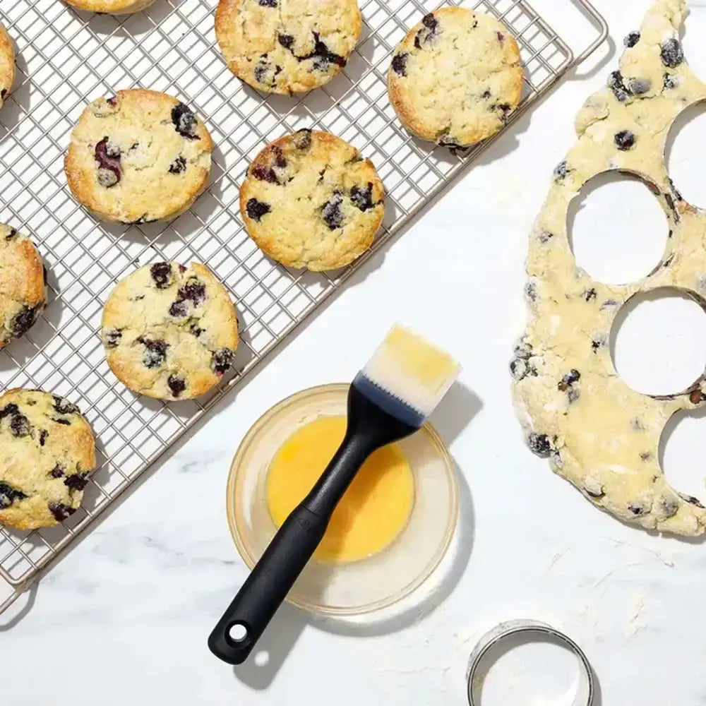 Blueberry muffins on a cooling rack with a brush and bowl of egg wash on a marble surface.