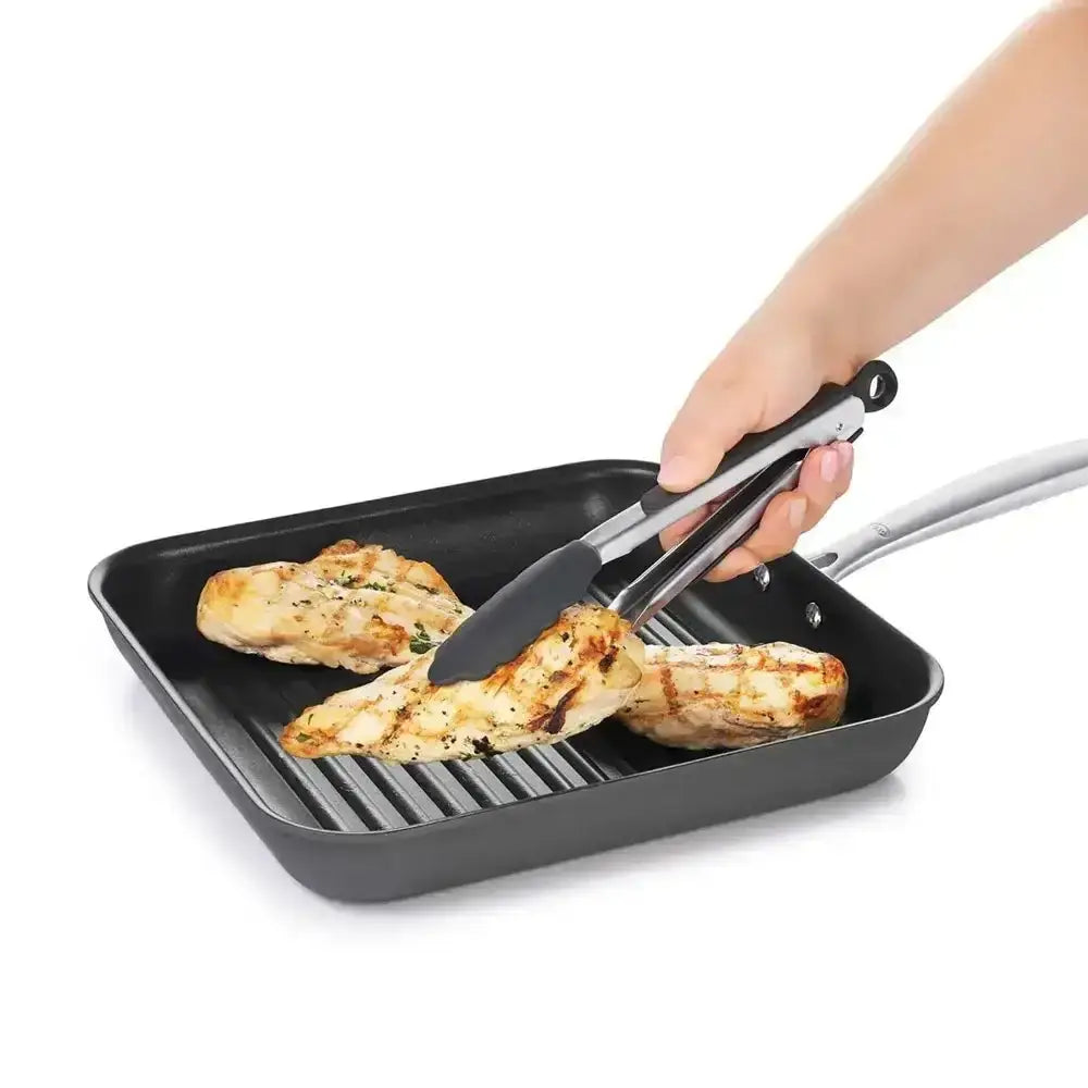 Person using tongs to handle grilled chicken in a black grill pan on a white background