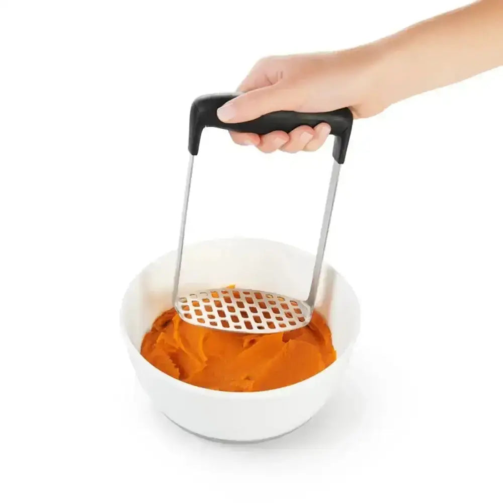 Hand using a potato masher to mash an orange potato in a white bowl on a white background