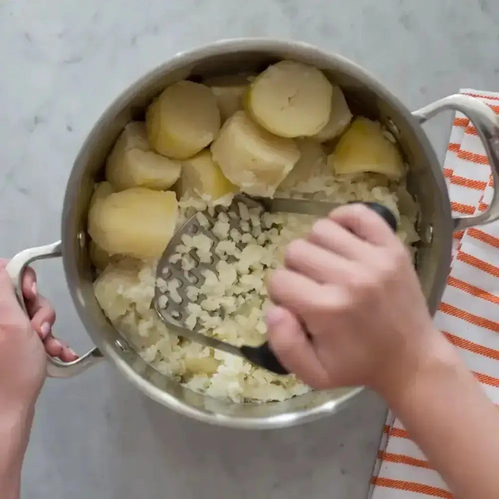 Person mashing potatoes in a pot with a masher on a light gray surface.